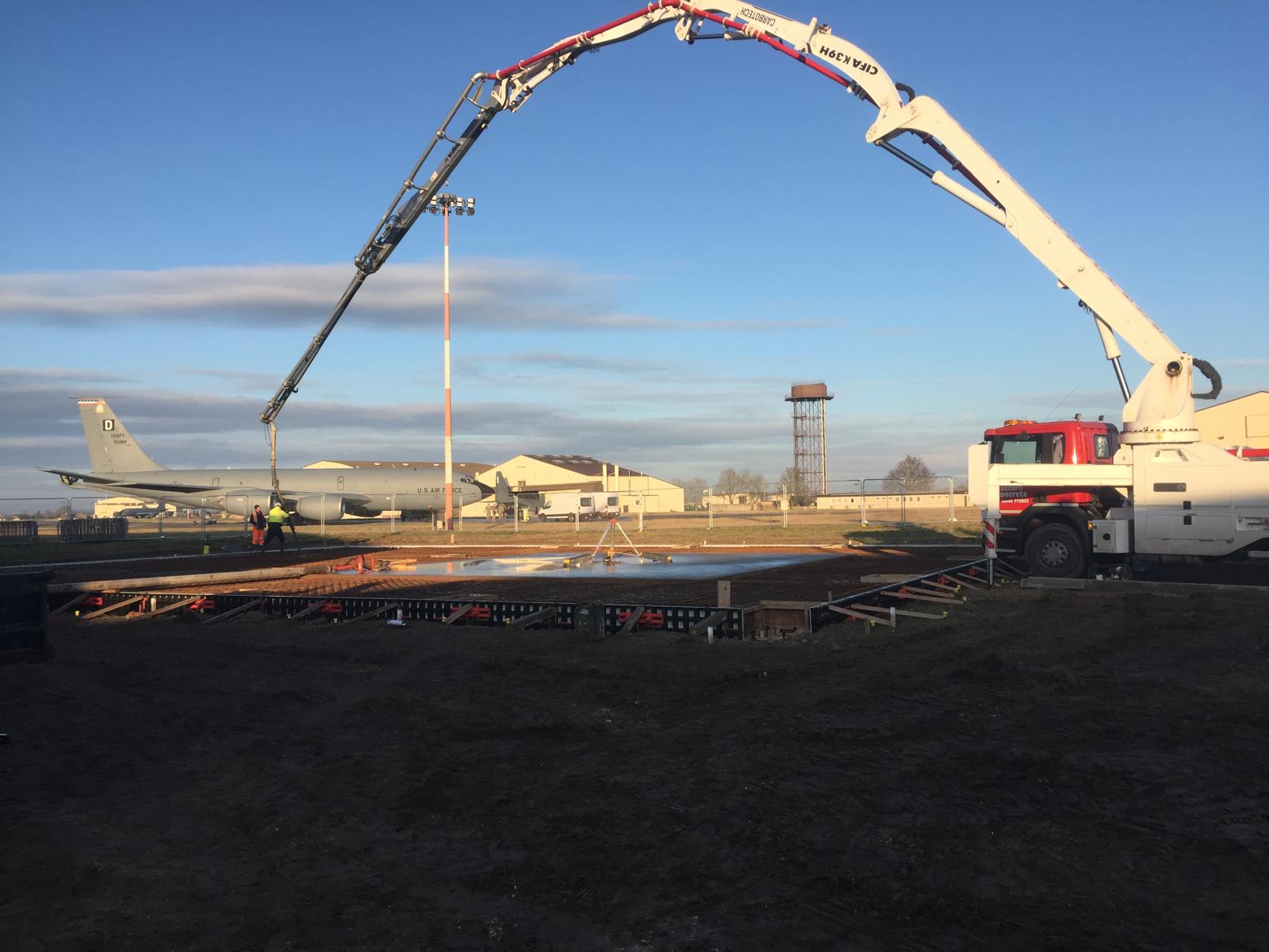 Concrete pour using a boom pump at RAF Mildenhall, with a US Air Force aircraft and airfield buildings beyond the works